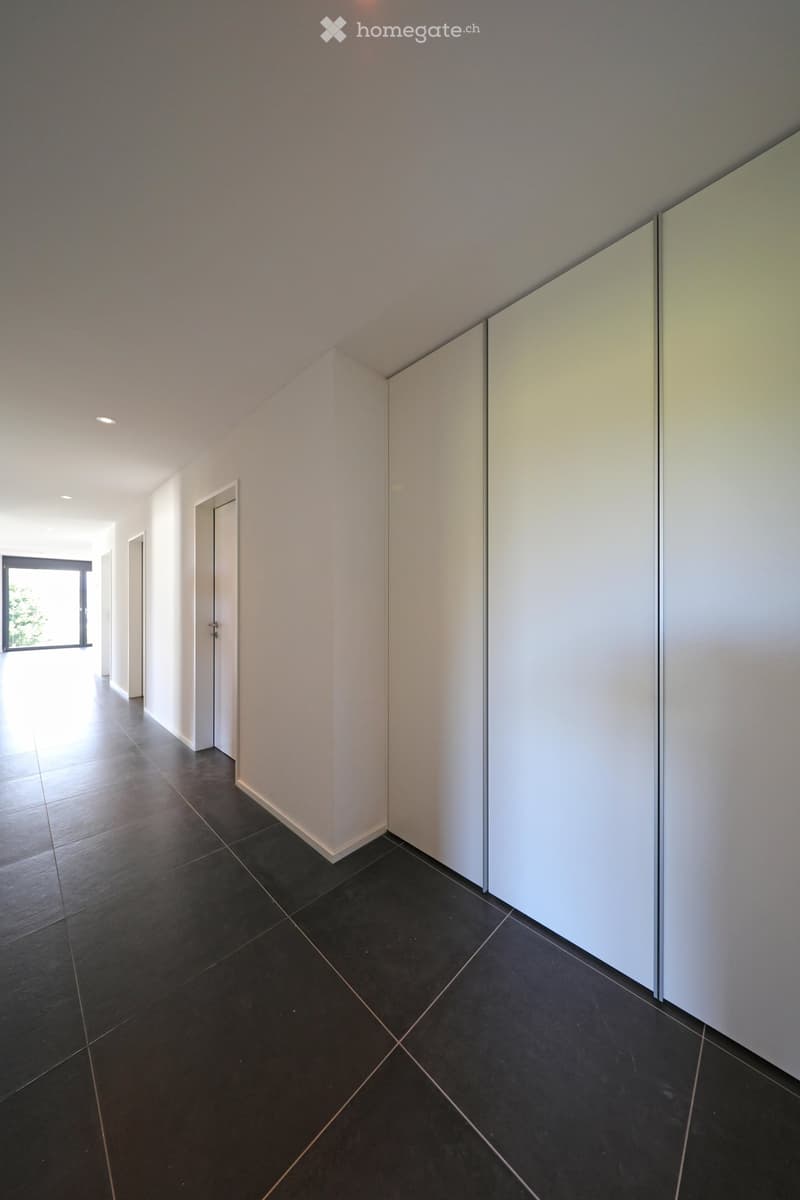 Empty hallway with dark tiled floor, white walls, and recessed lighting. Features a large window on the left and a closed door on the right.