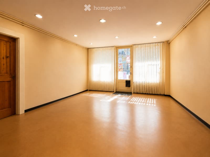 Empty hallway with wooden door, frosted glass door, and vertical blinds. Light-colored walls and floor, recessed ceiling lights.