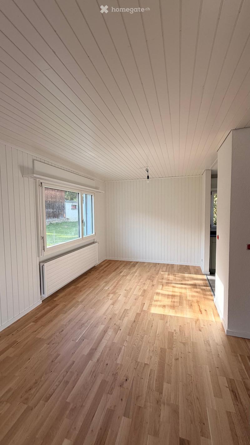 Empty room with wooden flooring, white paneled walls, and a slanted ceiling. Features a large window with a radiator below it, allowing natural light to enter.