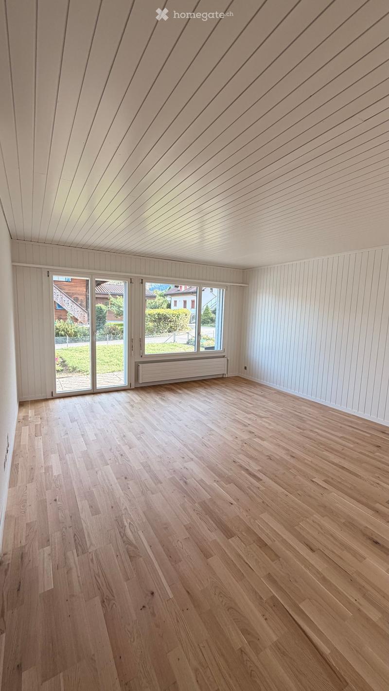 Empty room with light wooden flooring, white paneled walls, and a slanted wooden ceiling. Large windows and sliding doors lead to an outdoor area.