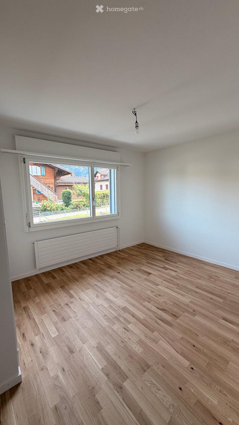 Empty room with light wooden flooring, white walls, large window with view of a house and garden, radiator, and a single ceiling light fixture.