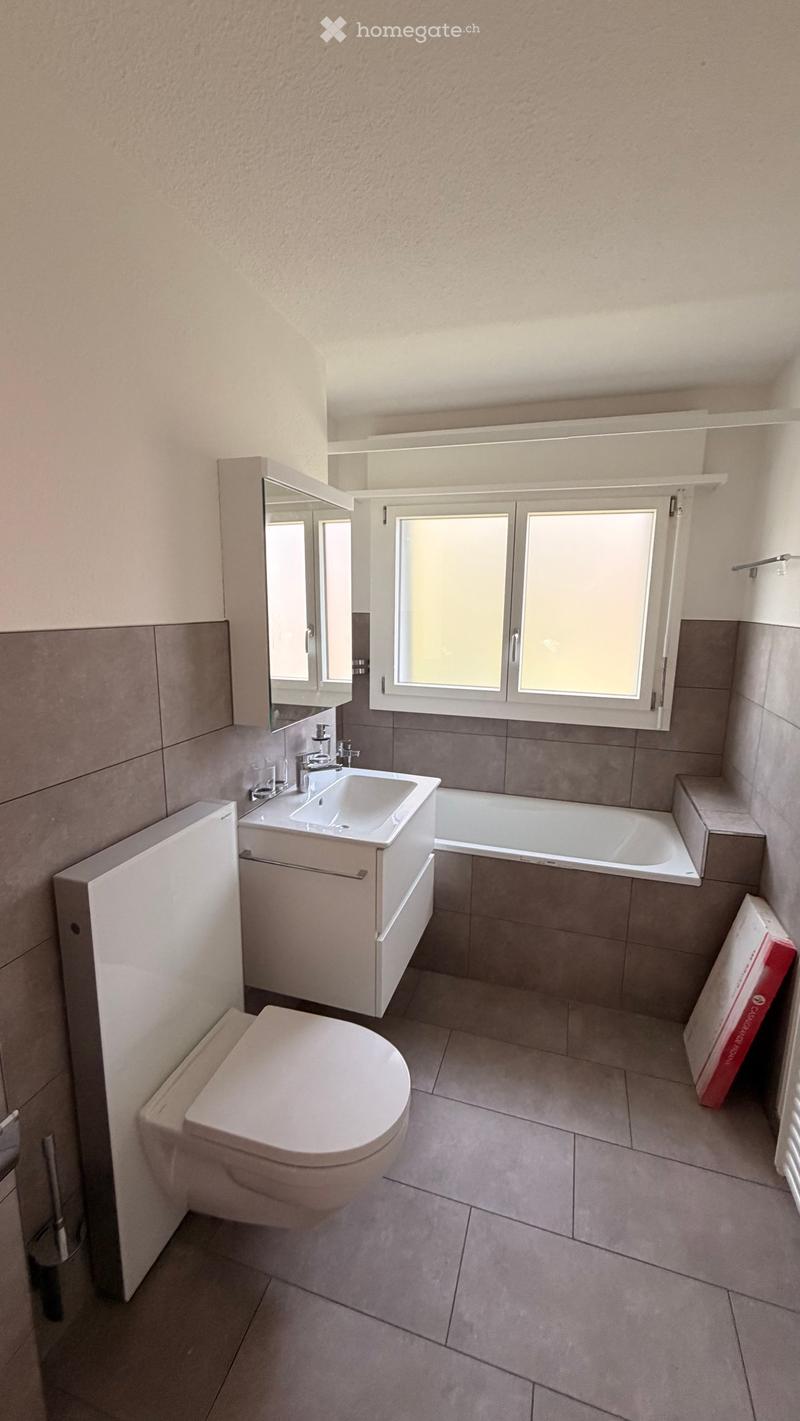 Modern bathroom with a toilet, sink, bathtub, mirror, and cabinet. Features tiled walls and a window with blinds.