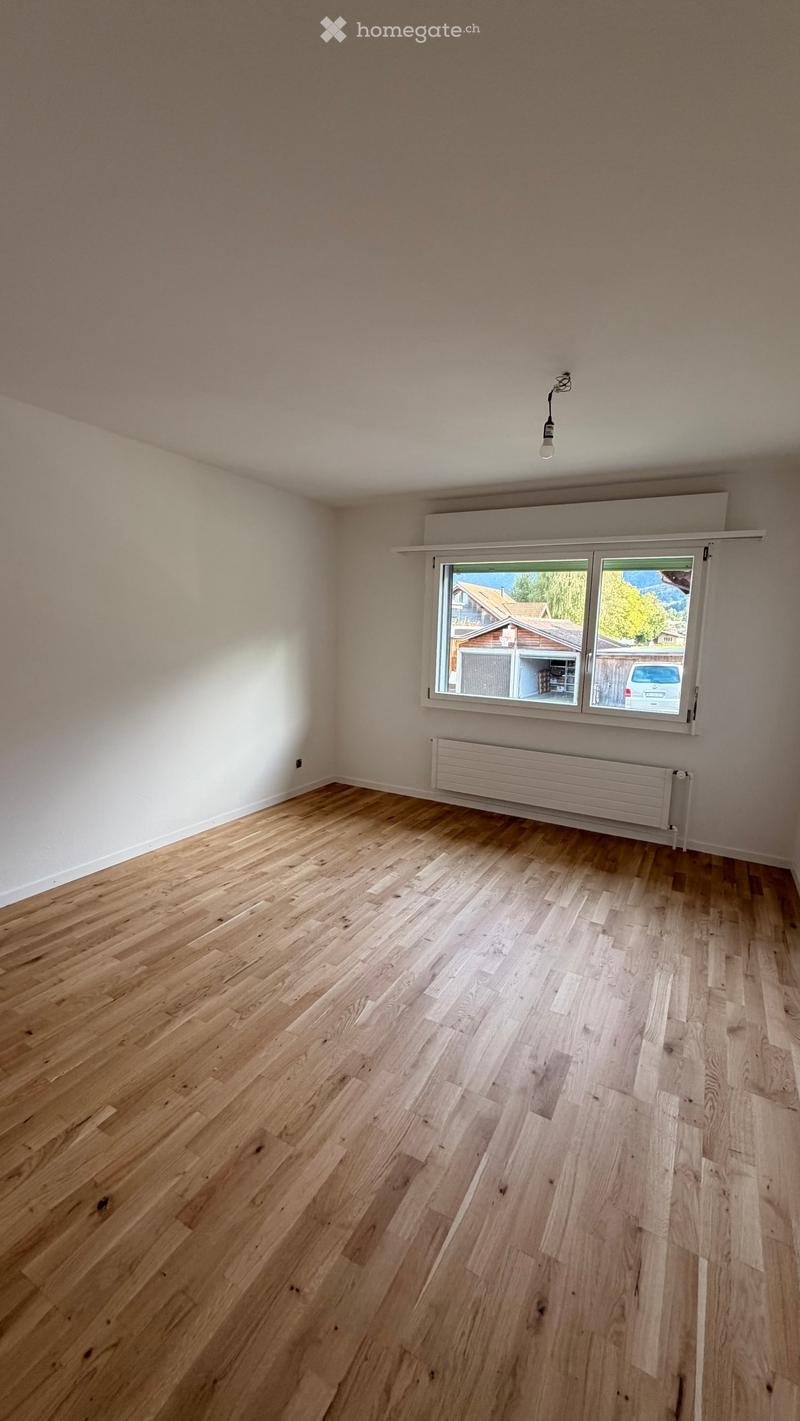 Empty room with light wooden flooring, white walls, a large window with a view of the outside, and a radiator below the window.