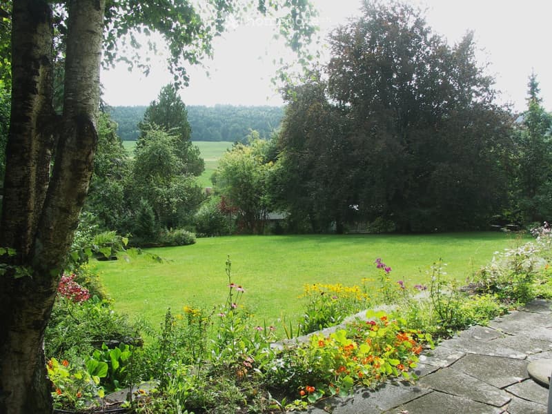 A well-maintained garden with a large, open lawn surrounded by trees and colorful flowers, featuring a stone pathway and a serene, natural setting.