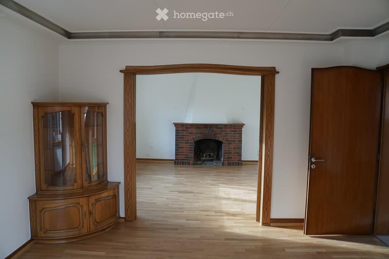 Empty hallway with wooden flooring, white walls, and a fireplace visible in the background.