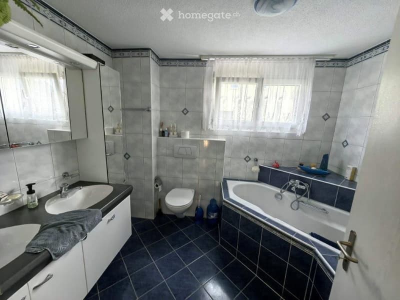 Bathroom with a sink, toilet, bathtub, and mirror. The room features tiled walls and a dark tiled floor, with a window covered by a white curtain. A towel is draped over the edge of the sink, and a small shelf holds various toiletries.