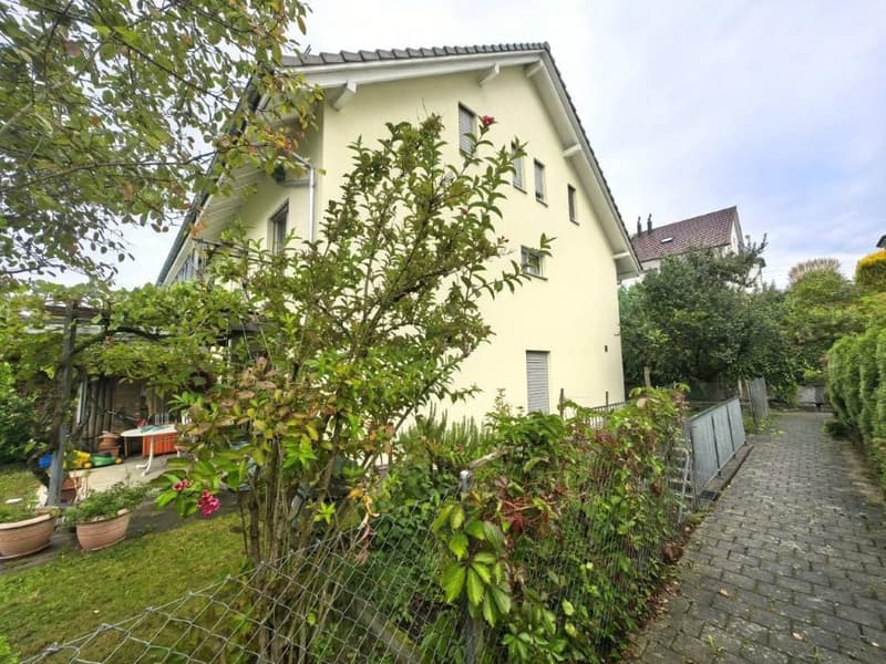A well-maintained garden area with a variety of plants and trees, a small table and chairs, and a pathway leading to a two-story house with a light yellow facade and multiple windows.