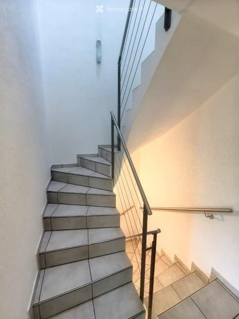 A modern staircase with gray tiled steps and a sleek metal railing, illuminated by natural light from a window at the top.