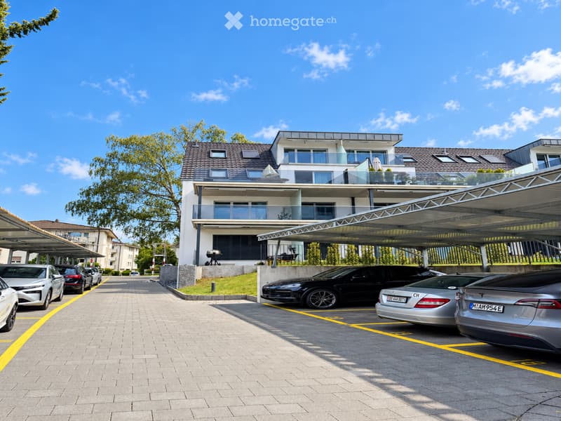 Modern multi-story residential building with balconies, surrounded by a paved parking area with several parked cars under covered structures.
