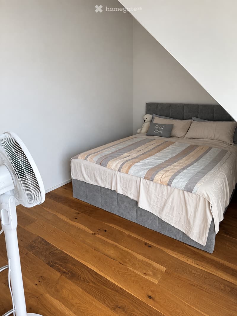 A cozy bedroom with a wooden floor, a large bed with a grey headboard, a striped duvet, pillows, and a small teddy bear toy. A white fan is positioned on the left side of the room.
