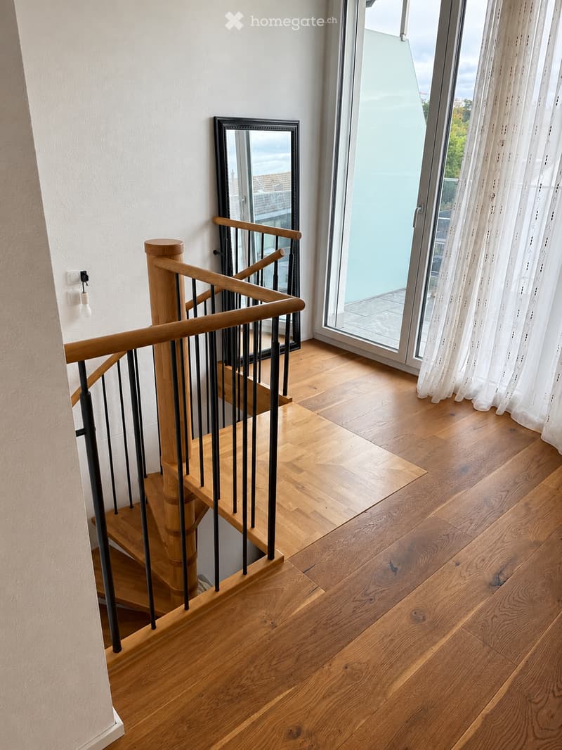 Spiral staircase with wooden steps and a black metal railing, leading up to a landing area with wooden flooring and a large window with white curtains.