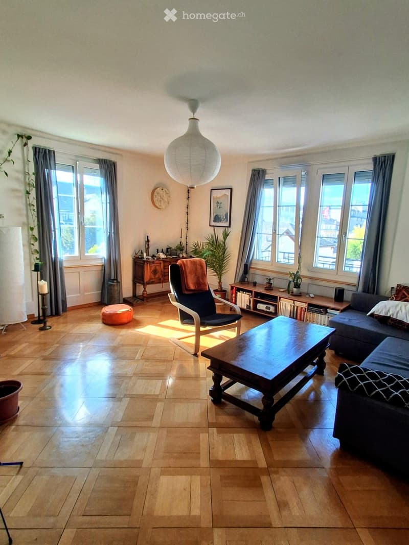 Bright living room with wooden flooring, large windows, a sofa, coffee table, armchair, and decorative plants.