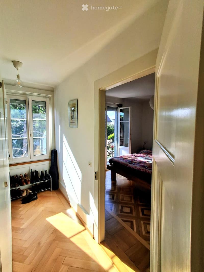 Bright hallway with wooden flooring, white walls, and a ceiling light fixture. A shoe rack with several pairs of shoes is visible on the left, and an open door leads to a bedroom with a bed and patterned bedding.