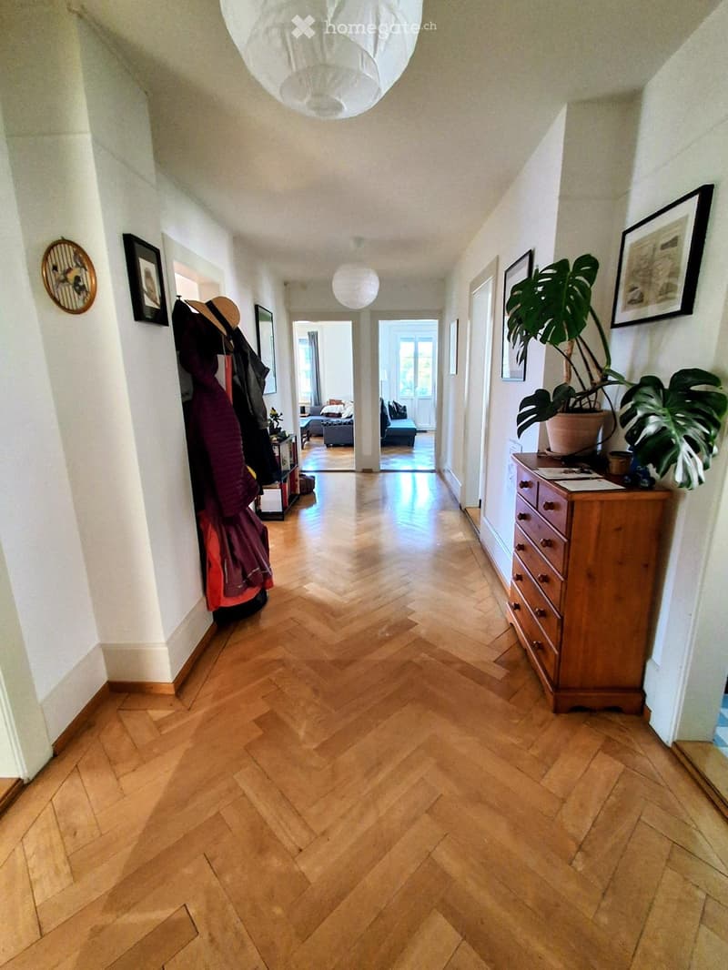 A long, well-lit hallway with herringbone wooden flooring, white walls, and several framed pictures. A coat rack with jackets and a potted plant are visible on either side, leading to adjacent rooms.