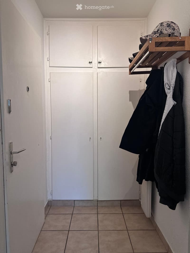 A narrow hallway with tiled flooring, white walls, and two white doors. A wooden coat rack with hooks holds several jackets and a hat on a shelf.