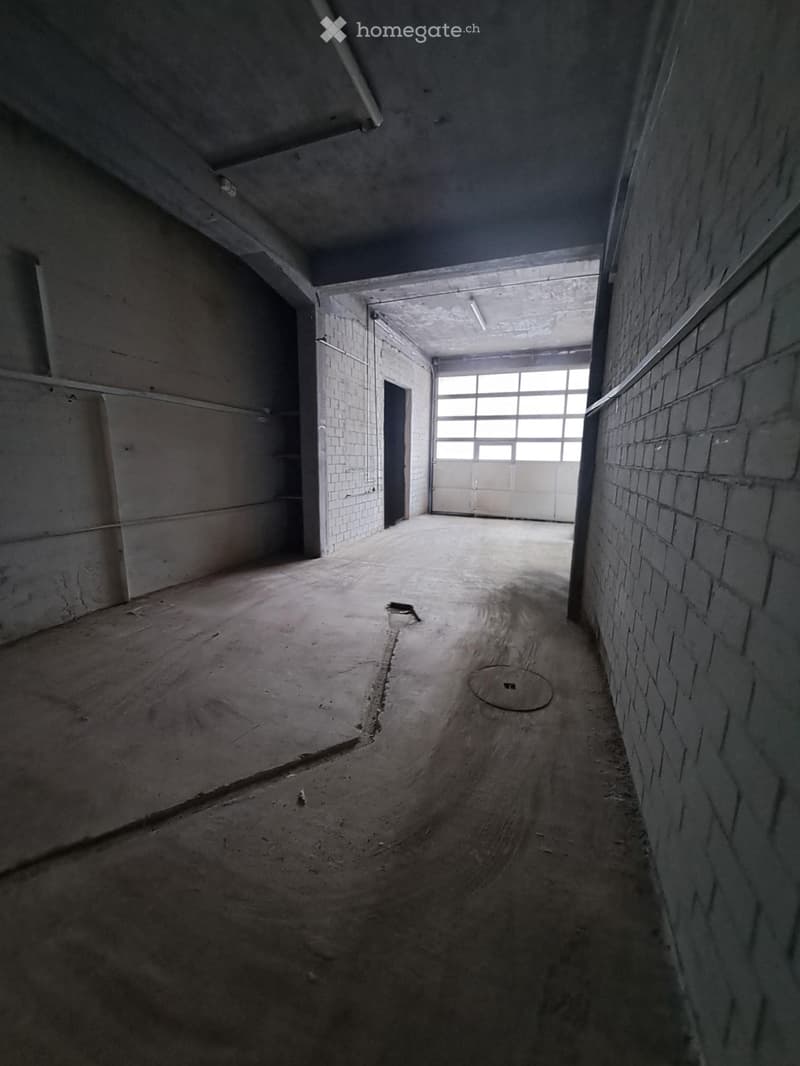 Empty industrial-style room with concrete floors, brick walls, and a large garage door at the far end.