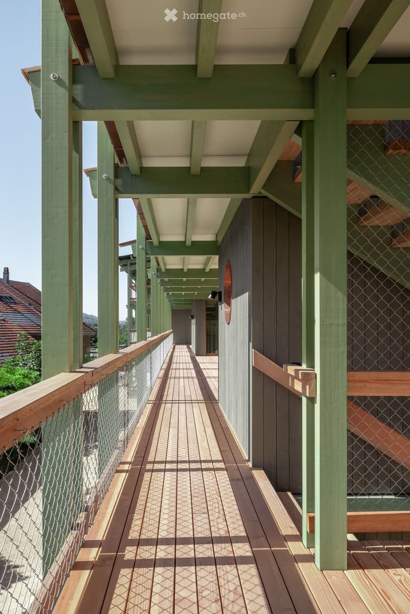 A wooden deck corridor with green railings, metal mesh fencing, and a tiled pathway leading to a building entrance.