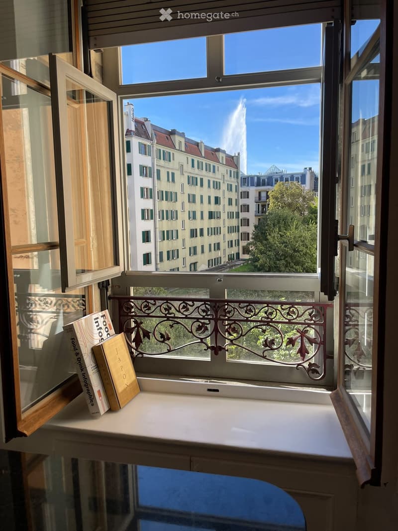 Balcony with ornate metal railing, overlooking cityscape with fountain and buildings, two books on the windowsill.