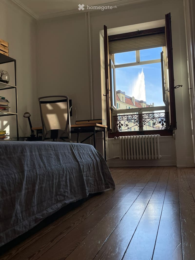 Bright bedroom with wooden flooring, large window offering a city view, bed with grey duvet, chair, small table, lamp, radiator, and shelf with books.