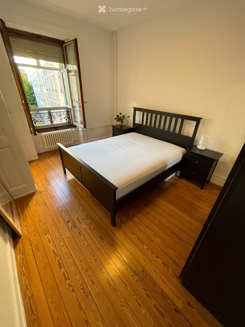 Cozy bedroom with wooden flooring, a double bed with a dark headboard, nightstands, a small plant, and a window with a view of the city.