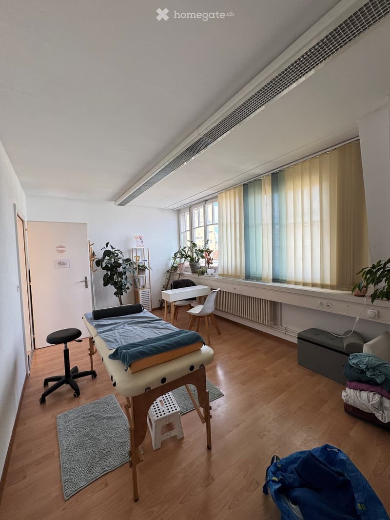 A well-lit room with wooden flooring, a massage table, a small table, chairs, a cabinet, and plants near the windows.
