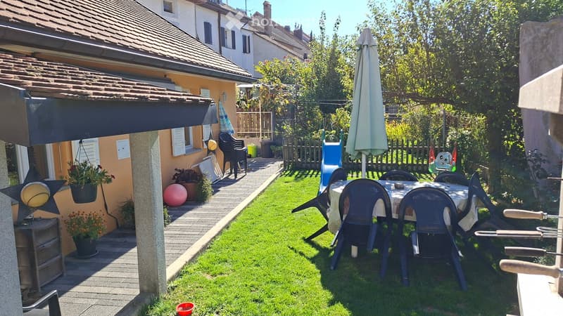 Garden with a dining table and chairs, a large umbrella, a grassy area, and a pathway leading to a house with a covered patio area.