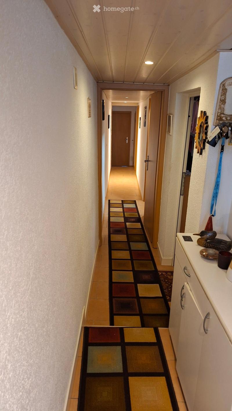 A long, narrow hallway with beige walls, wooden doors, and a colorful checkered rug leading to another section.