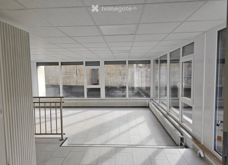 Empty room with white walls, tiled floor, large windows, and a metal railing, illuminated by natural light.