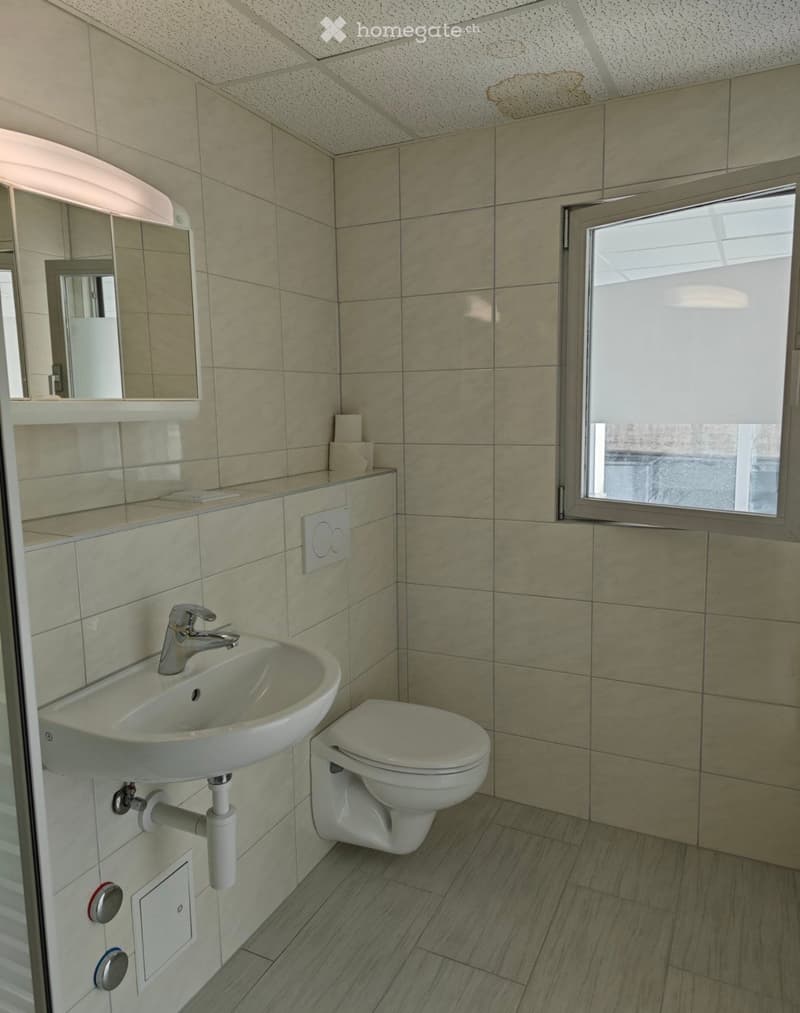 Modern bathroom with beige tiled walls, a white sink with a chrome faucet, a toilet, and a large mirror above the sink. The room features a window with blinds and a speckled ceiling.