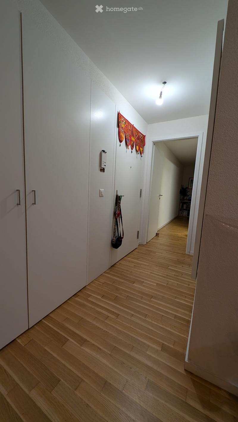 A narrow hallway with light wooden flooring, white walls, and a small decorative tapestry hanging on one wall. A door leads to another room, and a small shelf is visible in the background.