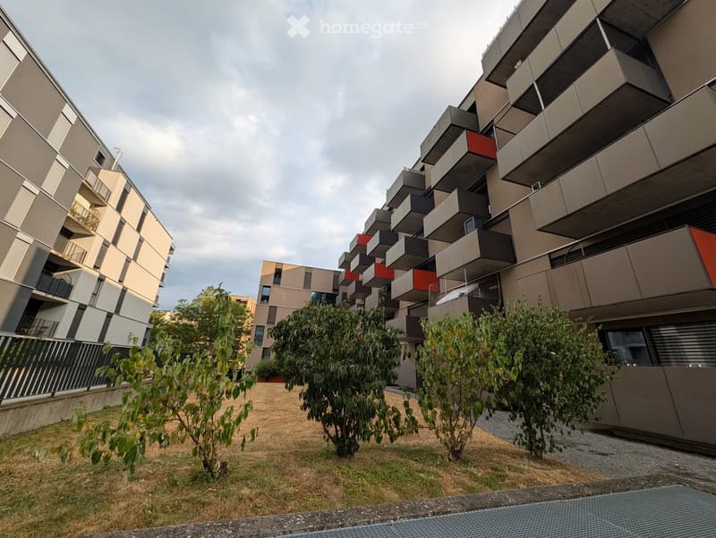 Modern multi-story residential building with a contemporary design, featuring balconies and greenery in the foreground.
