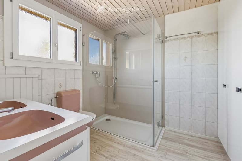 Modern bathroom with a glass-enclosed shower, a double sink, and a toilet. The space features light-colored tiles and wooden flooring.