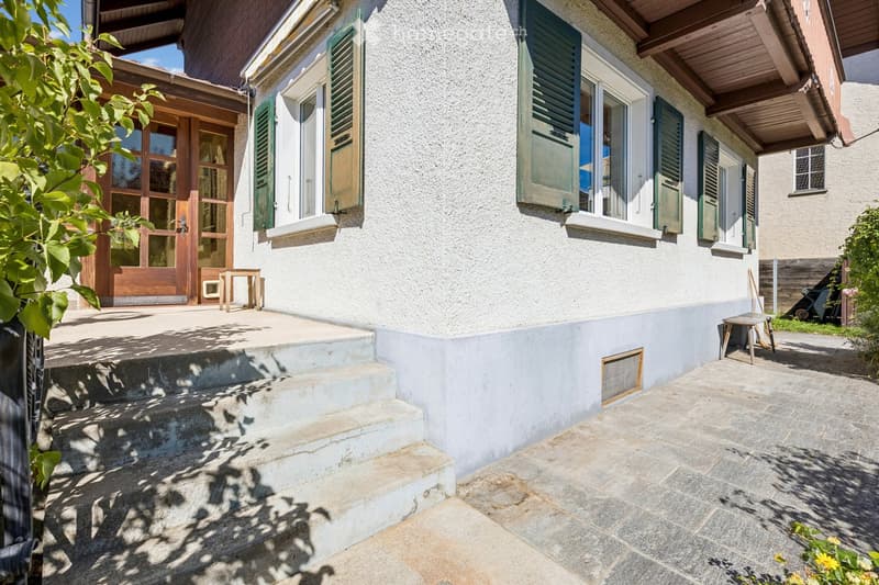 Two-story house with a raised ground floor, featuring a stone staircase leading to a wooden entrance door with glass panels. The facade includes green shutters and multiple windows, with a small patio area and outdoor furniture visible.