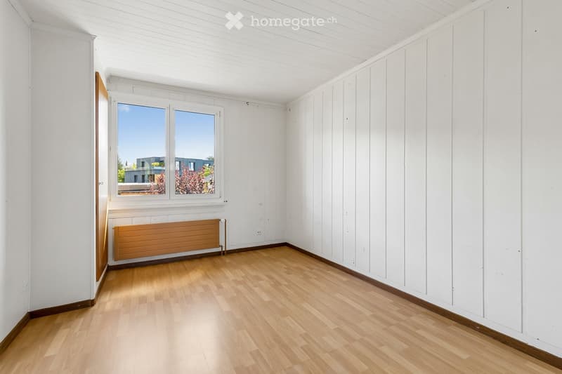 Empty room with wooden flooring, white paneled walls, and a large window offering a view of the outside. A radiator is visible below the window.