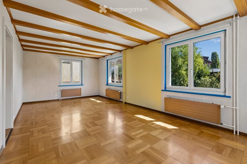 Empty room with wooden flooring, exposed wooden beams on the ceiling, and large windows with blue frames. The walls are painted in neutral tones, and there are radiators along the walls.