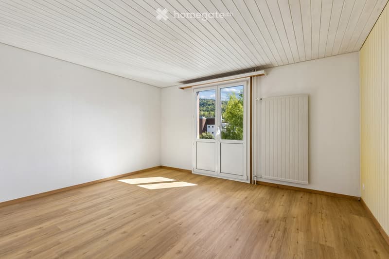 Empty room with wooden flooring, white walls, and a large window with a view of greenery outside.