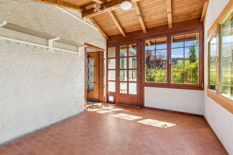 Bright hallway with terracotta tiled floor, wooden ceiling beams, large windows, and a wooden door leading to another room.