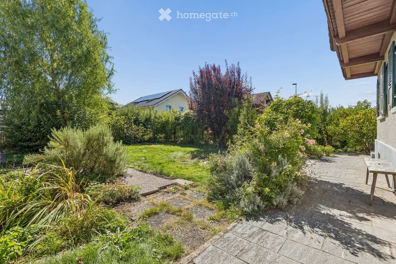 Well-maintained garden with a variety of plants, shrubs, and trees, featuring a paved pathway leading to a wooden bench near the house.