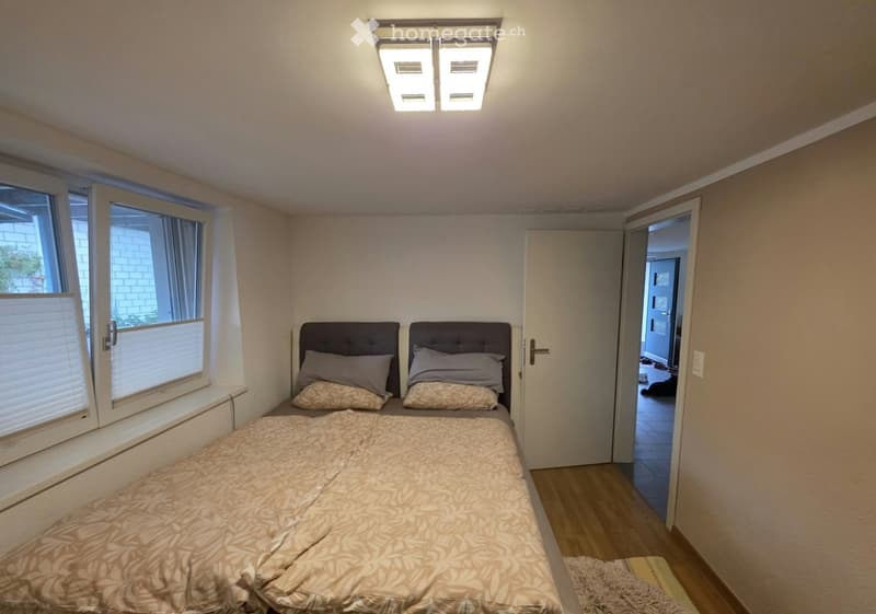 A small, well-lit bedroom with a double bed, headboard, pillows, and a window with blinds. The room features wooden flooring and a door leading to another area.