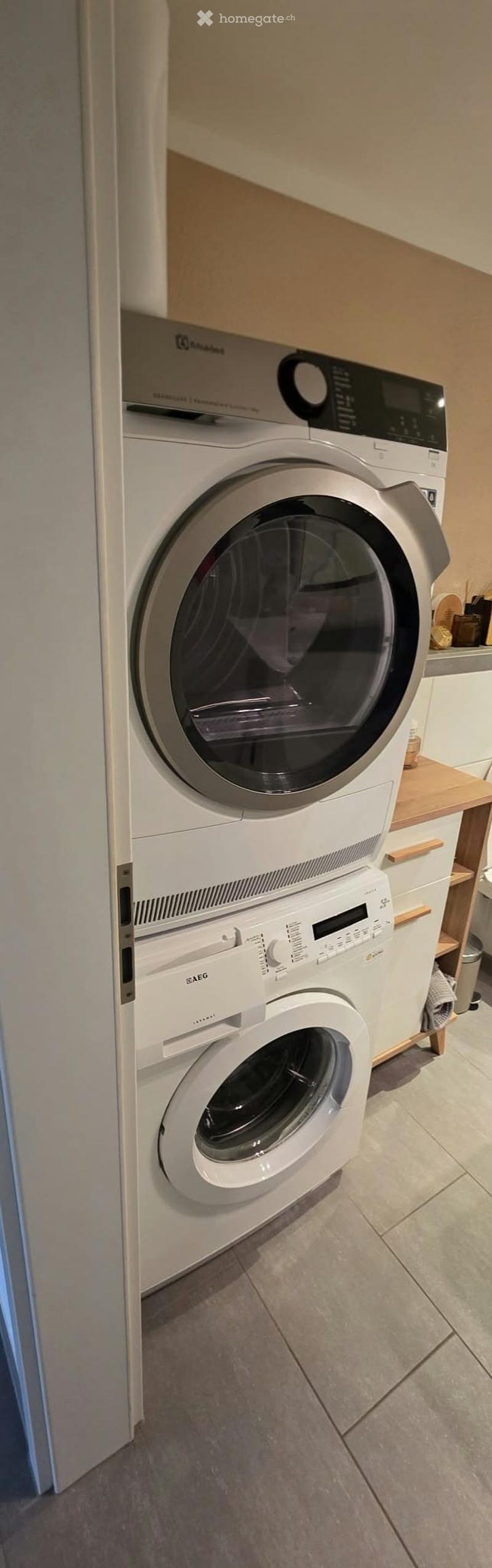 A modern bathroom with a white washing machine and a white dishwasher stacked vertically, with a wooden shelf and small items on the right side.