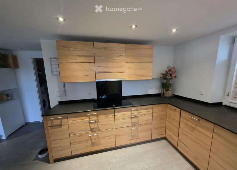 Modern kitchen with light wooden cabinets, black countertops, and stainless steel appliances. Features include a built-in oven, a vase with flowers on the counter, and recessed ceiling lighting.