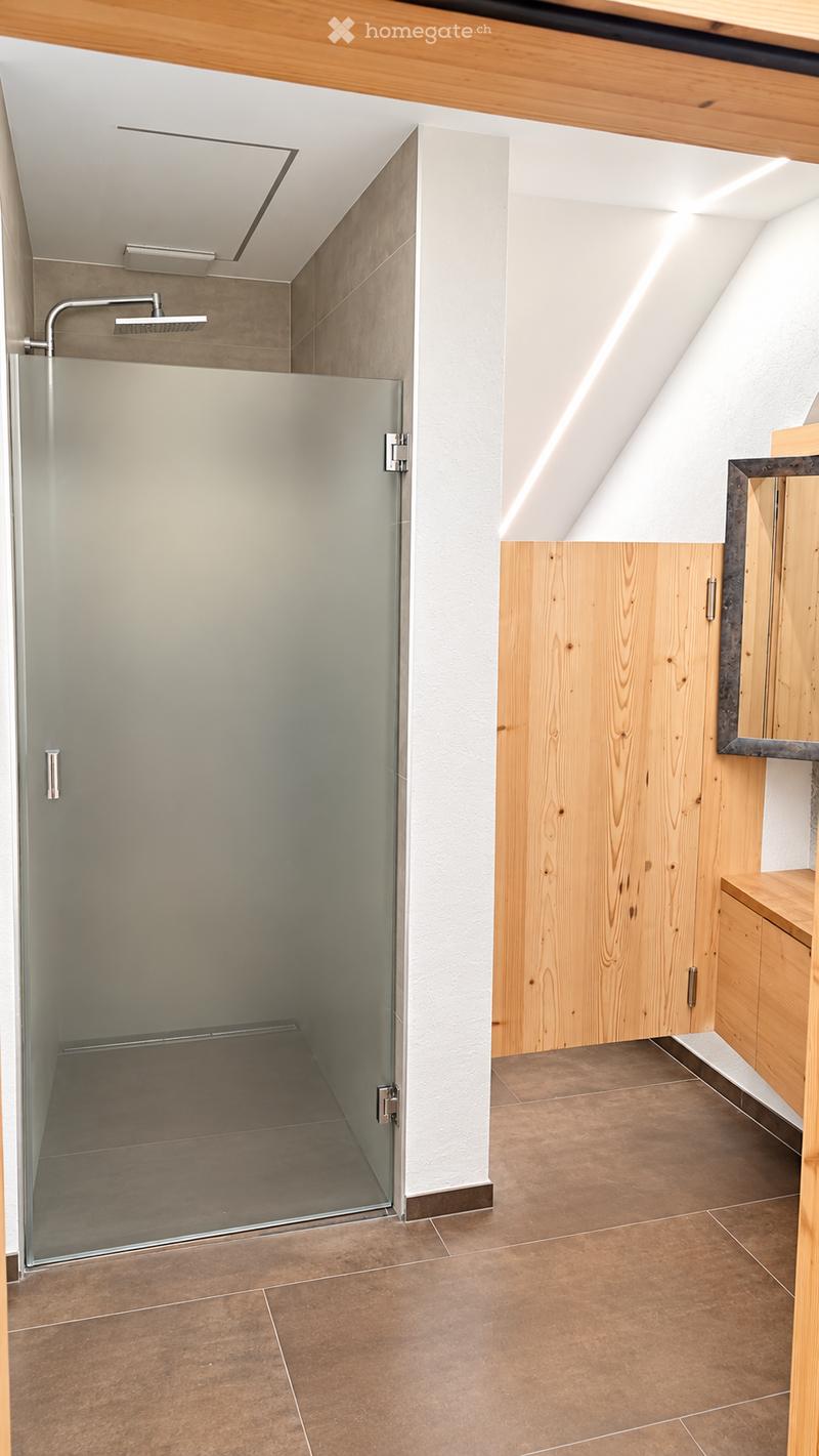 Modern bathroom with a glass-enclosed shower, wooden cabinetry, and a mirror. The space features a sloped ceiling and warm-toned tiles.
