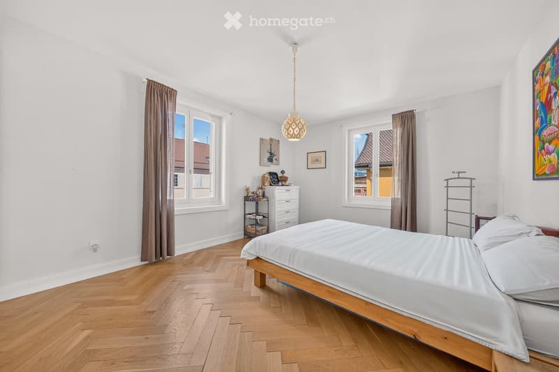 Bright bedroom with wooden flooring, large windows with brown curtains, a bed with white linens, a nightstand, and decorative items on shelves.