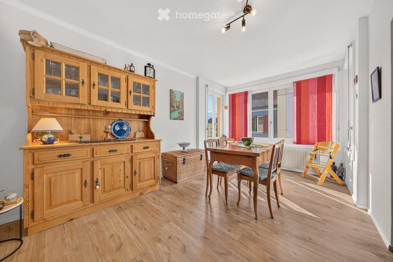 Bright, wooden-floored room with a large wooden cabinet, a dining table with chairs, a lamp, and decorative items including a painting and a wicker basket.