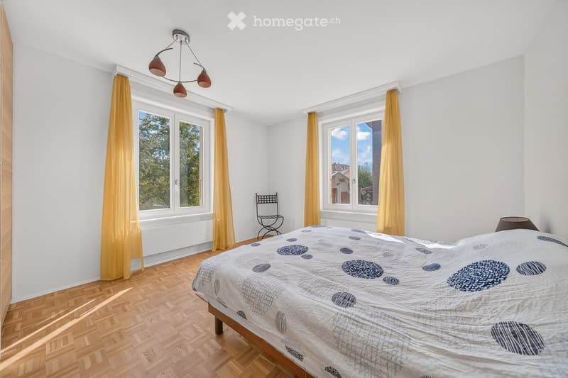 Bright bedroom with wooden flooring, large windows with yellow curtains, a bed with a patterned duvet, a small black chair, and a ceiling light fixture.