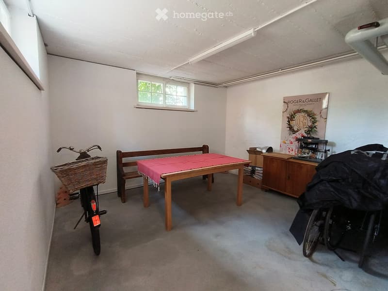 A small, furnished room with a wooden table covered in a red cloth, a bench, a bicycle, a cabinet, and a poster on the wall.