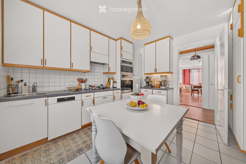 Modern kitchen with white cabinets, a central island table, and various appliances including a dishwasher, oven, and refrigerator. The island features a bowl of fruit and a coffee maker on the counter.