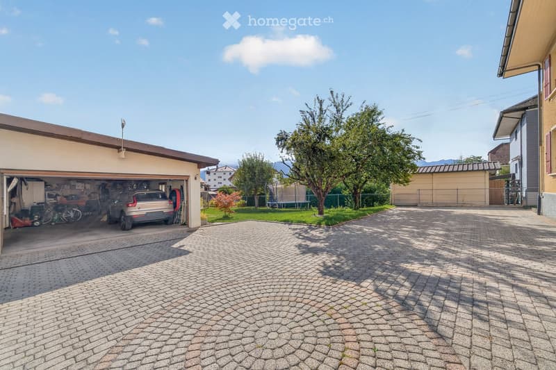 Driveway leading to a garage with a parked car, surrounded by greenery and neighboring houses.