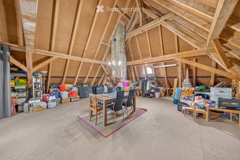 Attic space with exposed wooden beams, concrete floor, and various stored items including a dining table, chairs, shelves, and clothing racks.