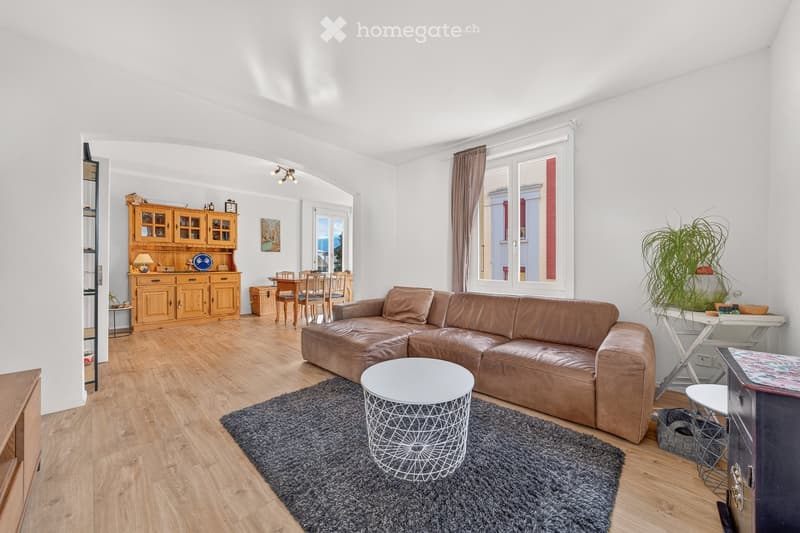 Bright living room with a brown leather sofa, white coffee table, wooden cabinet, dining area with table and chairs, and a potted plant near the window.